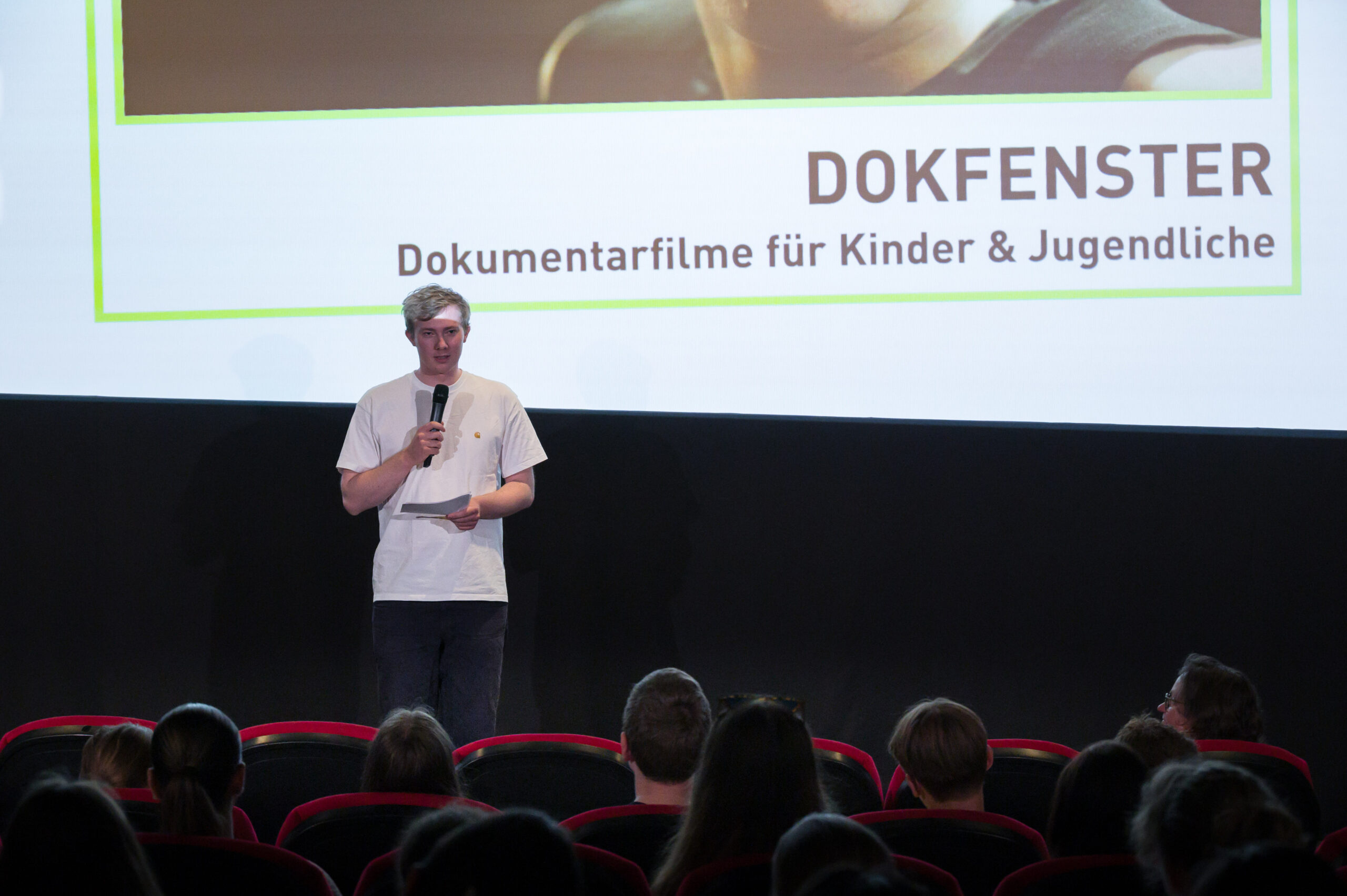 Jakob Michal standing in a cinema in front of an audience of school children, presenting his short documentary ‘Tough Moves,’ with a film still projected on the screen behind him.